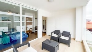 Een balkon met rieten meubilair en een glazen deur die leidt naar een slaapkamer met een bed en bureau.