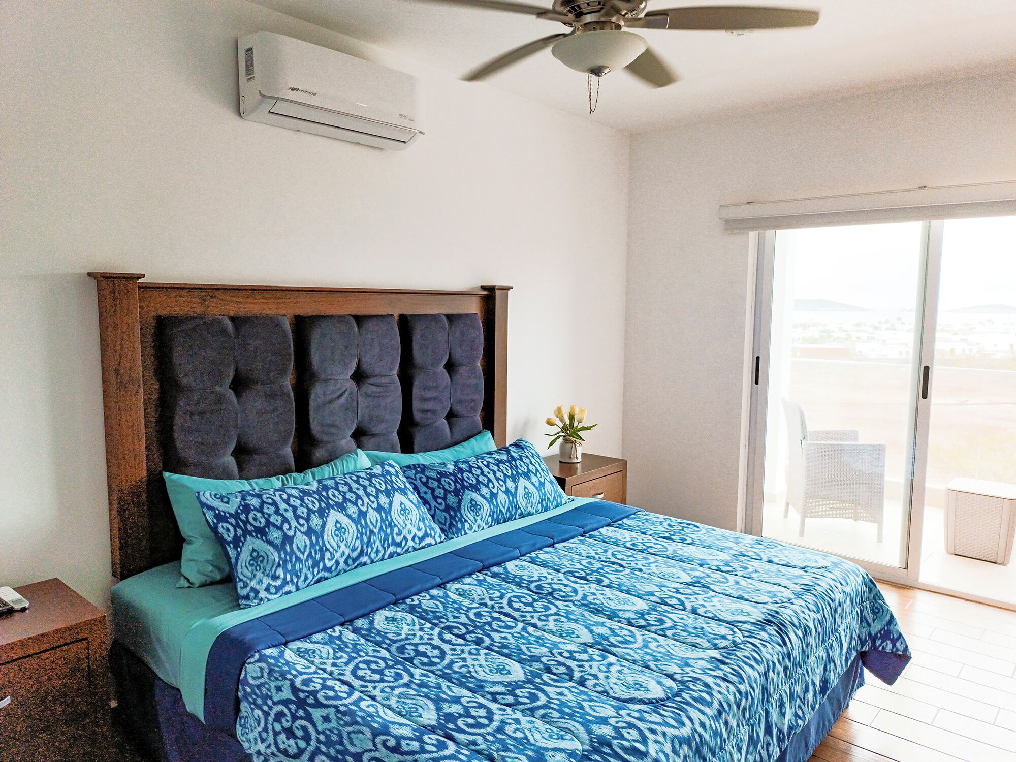 A bedroom with a large bed, a ceiling fan, an air conditioning unit, a nightstand, a small table with a vase of flowers, and a sliding glass door to a balcony.