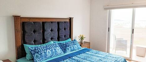 A bedroom with a large bed, a ceiling fan, an air conditioning unit, a nightstand, a small table with a vase of flowers, and a sliding glass door to a balcony.