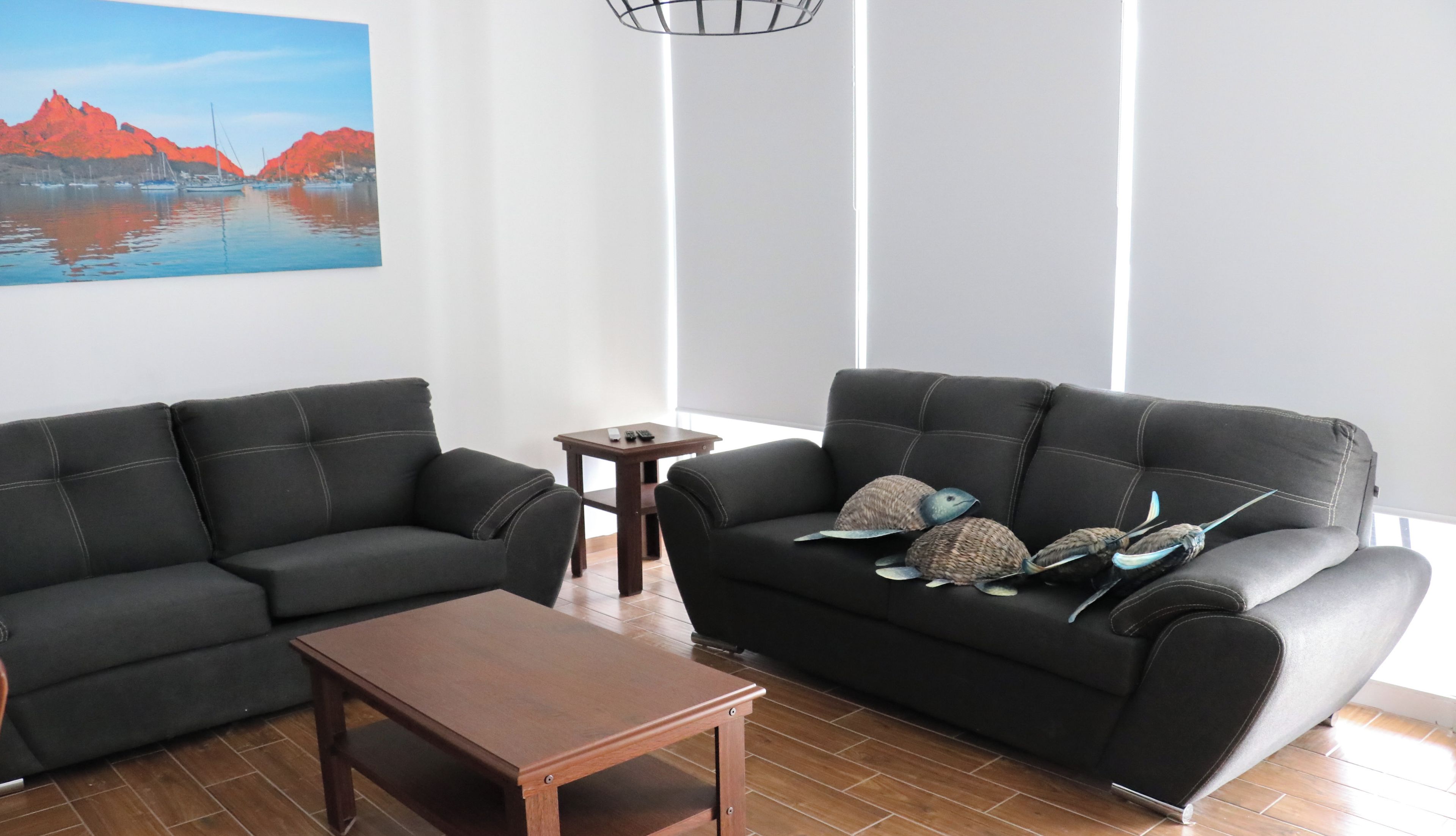 A living room with two black sofas, a wooden coffee table, and a small side table. A painting of a coastal scene is on the wall.