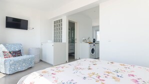 A bedroom with a bed, a TV, a patterned chair, a washing machine, and a kitchen area. - YourHouse Monges sea view-studio (Can Picafort)