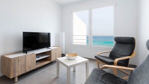Una sala de estar moderna con un televisor de pantalla plana, un mueble de entretenimiento de madera, una mesa de centro blanca, un sofá gris y vistas al océano a través de amplios ventanales.