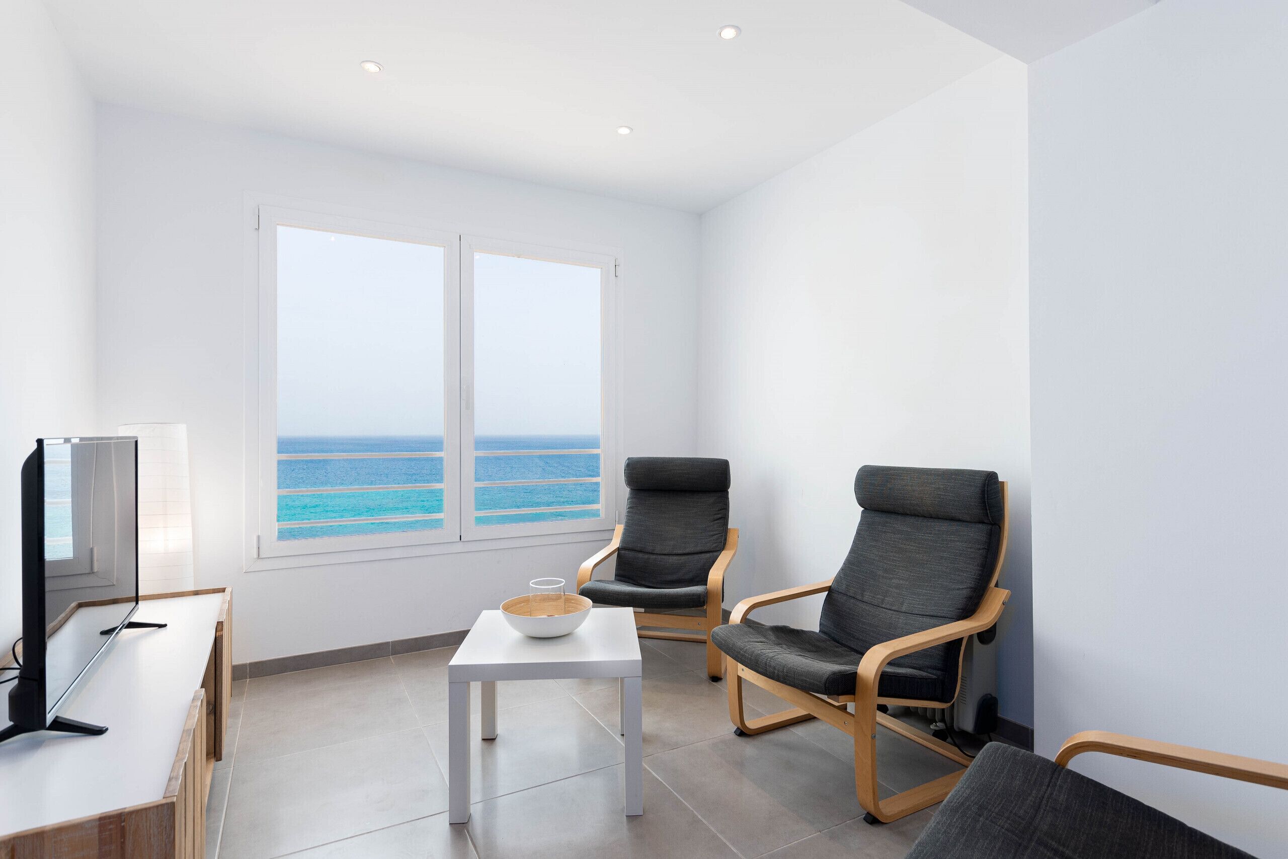 A modern room with a TV, two grey chairs, a small table with a bowl, and a large window with a view of the ocean.