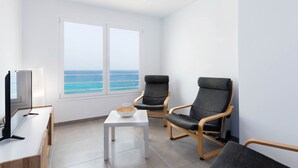 Una habitación moderna con televisor, dos sillas grises, una mesita con un cuenco y un ventanal con vistas al océano.