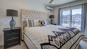 A bedroom with a tufted headboard, a large window with curtains, a ceiling fan, and a nightstand with a lamp. - The Vue #25 | Luxury Poolside Townhouse (Saint George)