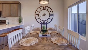 A dining area with a wooden table, white chairs, woven placemats, and a large wall clock. - The Vue #25 | Luxury Poolside Townhouse (Saint George)