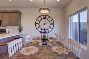 A dining area with a wooden table, white chairs, woven placemats, and a large wall clock. - The Vue #25 | Luxury Poolside Townhouse (Saint George)