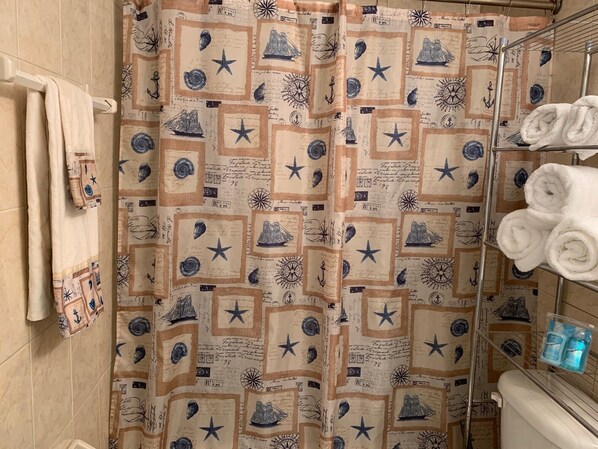 A bathroom with a nautical-themed shower curtain, a towel rack with folded towels, and a shelf with toiletries. - DAYTONA GETAWAY OCEAN FRONT CONDO (Daytona Beach)