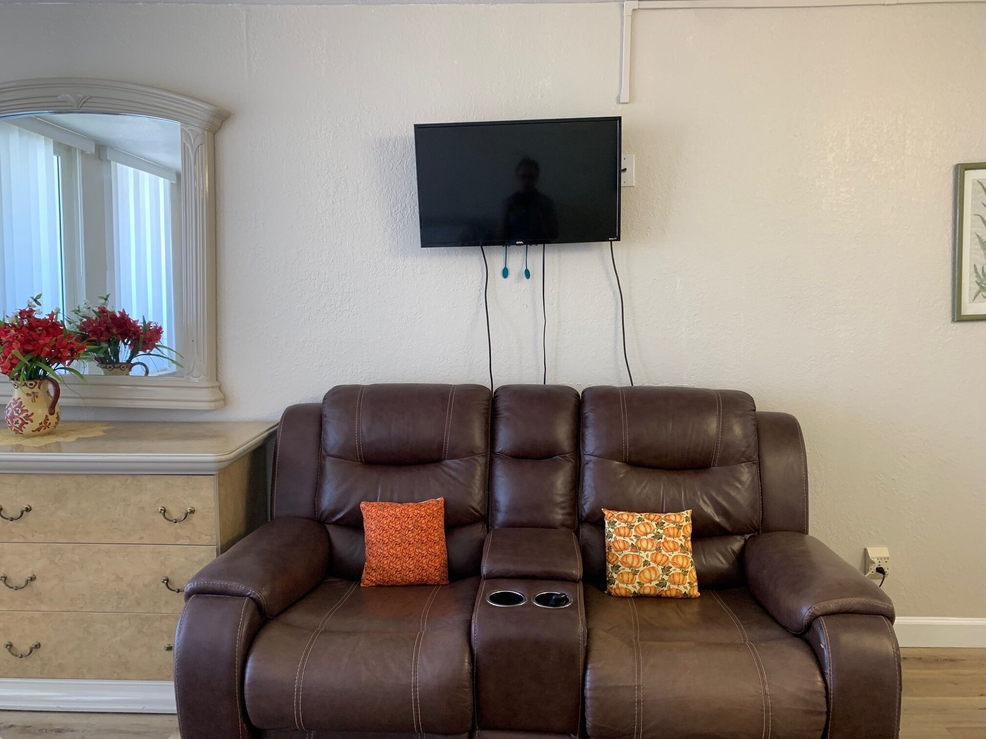 Een bruine leren bank met twee decoratieve kussens, een aan de muur bevestigde televisie, en een raam met jaloezieën.