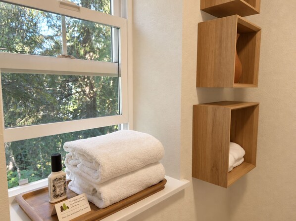 A window with a view of trees, a stack of white towels, a bottle of hand soap, and wooden shelves.