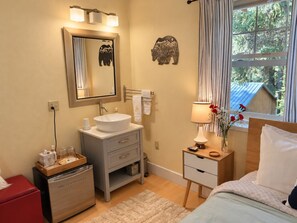 A hotel room with a large window, a bed, a bedside table with a lamp, a wooden nightstand, a bathroom with a sink, mirror, and towel rack.
