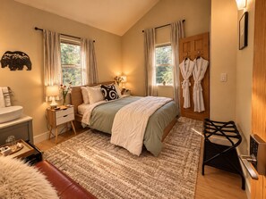 A bedroom with a bed, bedside tables, a nightstand, a window with curtains, a wooden door, and a robe rack.