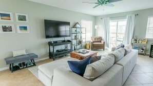 A living room with a large sofa, a coffee table, a TV, and a bookshelf. - Live the Salt Life in Golden Dog's Destin Rental (Destin)