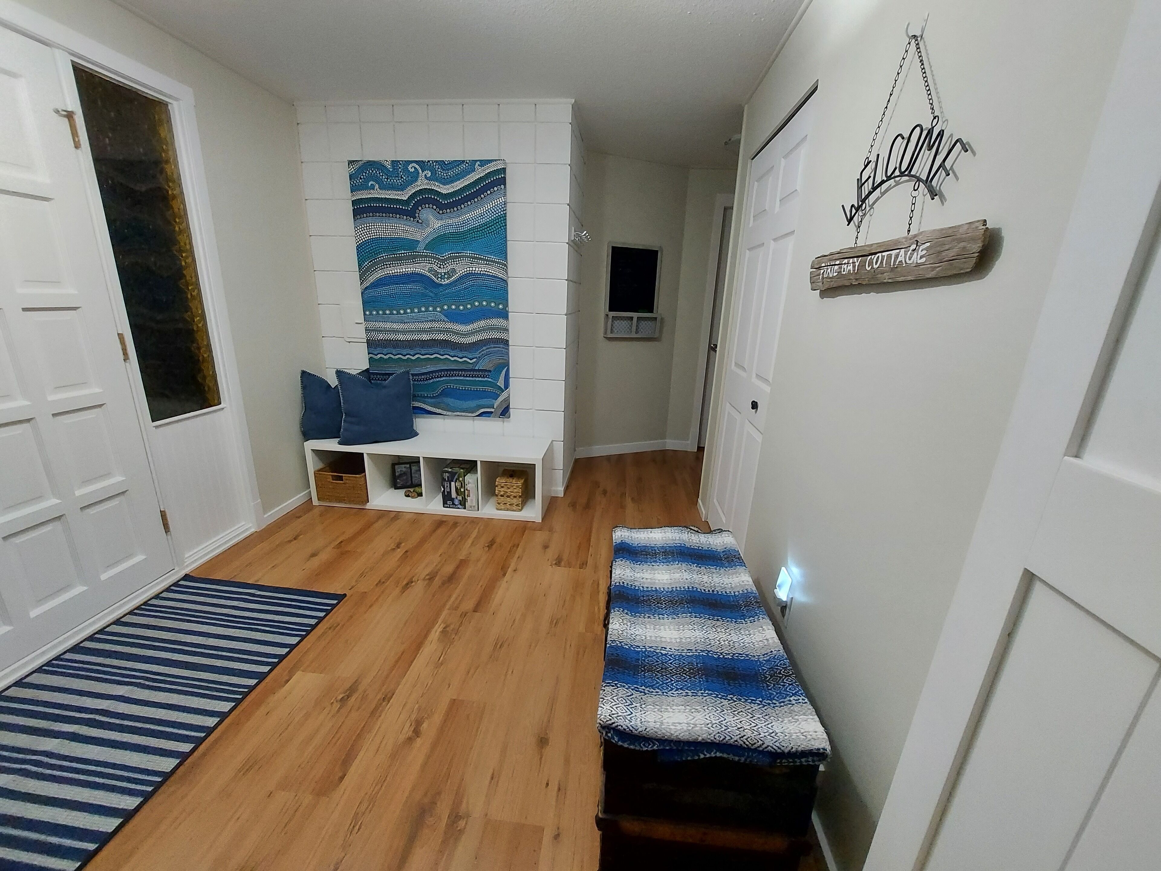 A hallway with a wooden floor, a bench, a wall-mounted welcome sign, and a patterned tapestry.