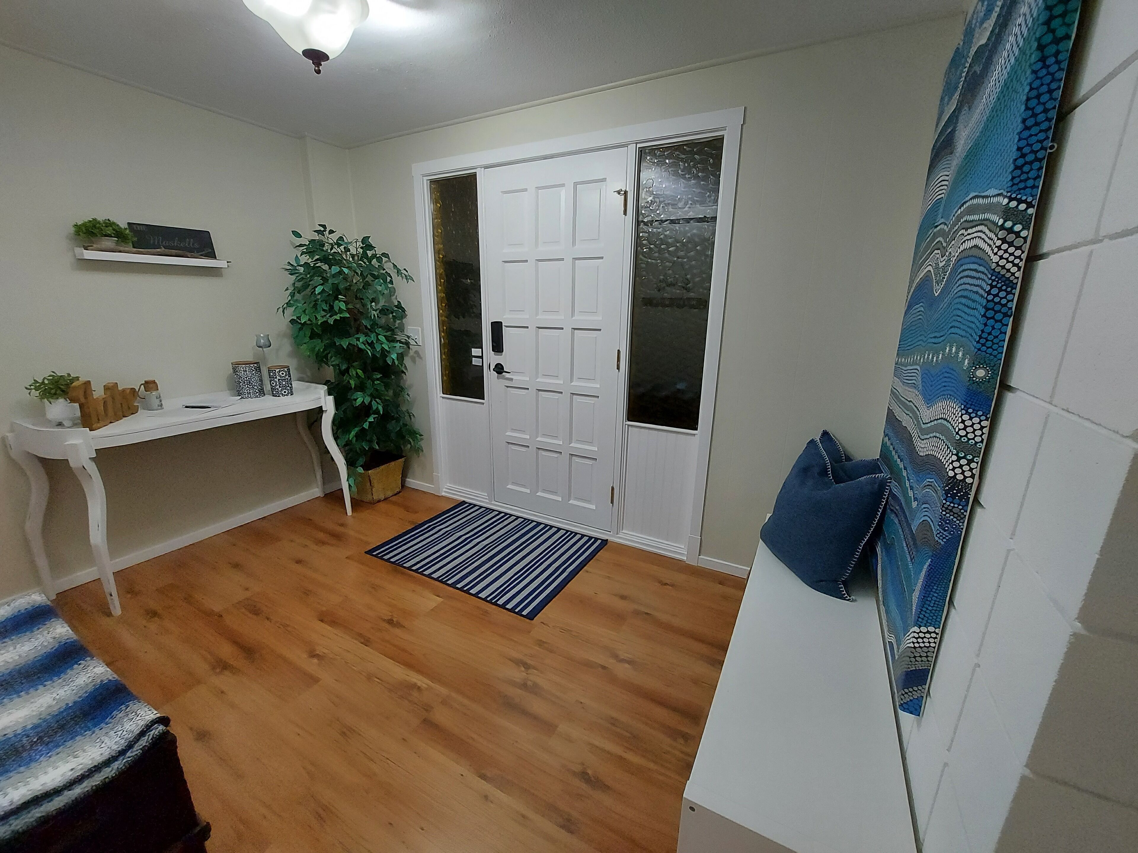 A room with a wooden floor, a white door with frosted glass panels, a white desk with a plant, and a blue patterned wall hanging.