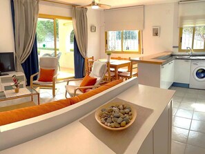 A modern living room with a sofa, chairs, a dining table, a TV, and a kitchen area with a washing machine.