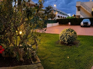 A well-maintained garden with a small bush and a tree with red flowers, a paved pathway, and a building with a staircase and a car parked in front.
