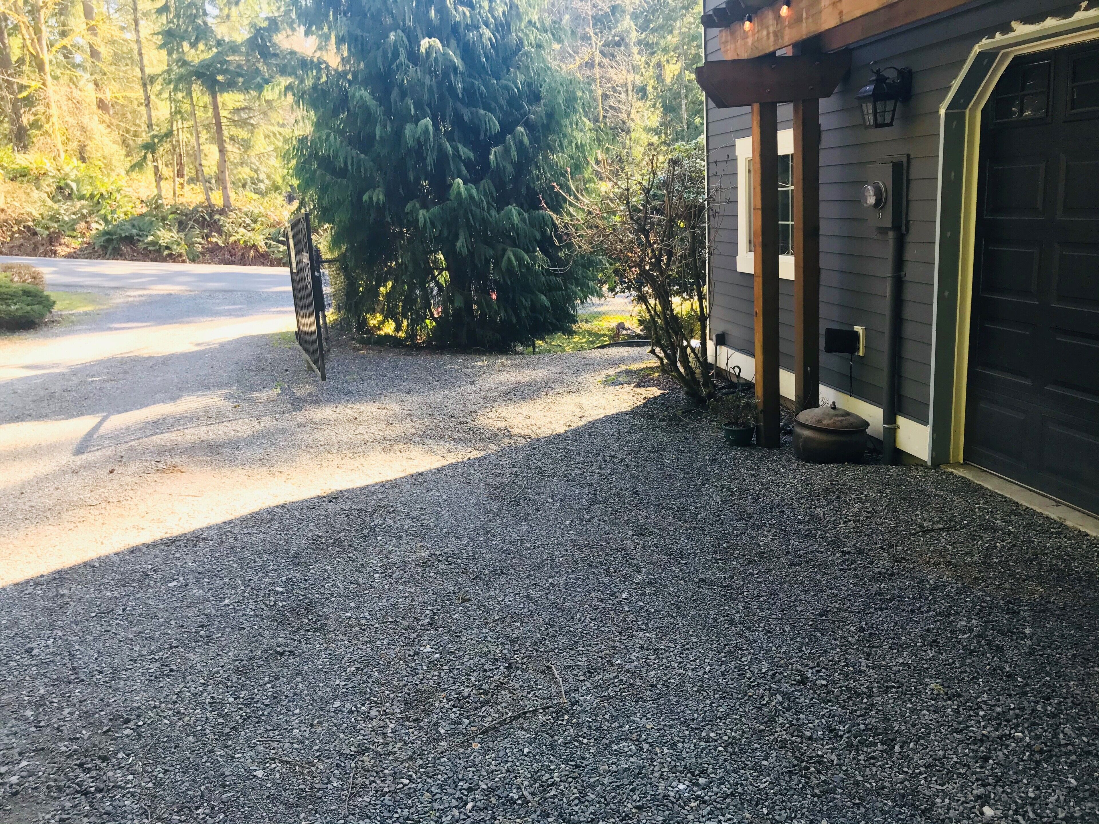 En grusvej fører til et hus med en garageport, omgivet af træer og en busk.