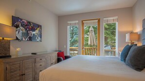 Ein Schlafzimmer mit einem großen Bett, einem an der Wand montierten Fernseher, einer hölzernen Kommode und einem Balkon mit Blick auf Bäume.