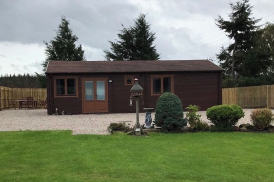 A single-story wooden building with a brown door and windows, surrounded by a gravel driveway and a small lawn with a trimmed bush.