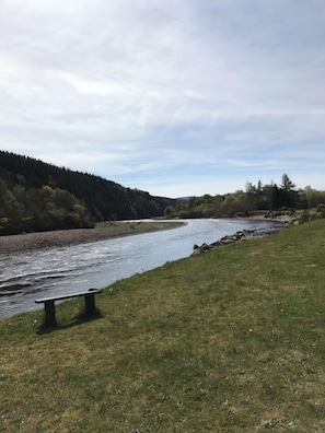 A river with a bench on the grassy bank, surrounded by trees and hills.