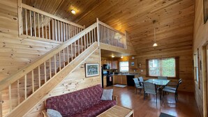 A wooden cabin interior with a staircase, a sofa, a dining table, and a kitchen area. - Heron Lodge - The Lodges at Oak Point, 1000 Islands - Right on the River! (Hammond)