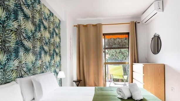 Une chambre d’hôtel avec un lit, une couvre-lit verte, une fenêtre avec des rideaux, une chaise placée à l’extérieur de la fenêtre et une commode en bois.