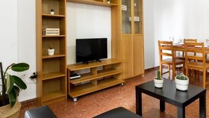 Un salon avec un téléviseur posé sur un meuble en bois, une table à manger avec des chaises et une grande bibliothèque en bois.