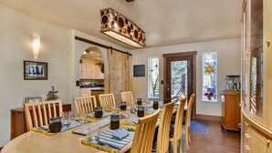 A dining area with a long wooden table set for six, surrounded by wooden chairs. There is a kitchen visible through an archway, and a large window with a star decoration. - Mt.Benson Suite at Bell In the Woods B&B (King/Queen/2xTwin/Bottom Flr) Max6ppl (Seward)