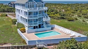 A multi-story building with a pool and outdoor seating, surrounded by greenery and overlooking a body of water. - Premier Oceanfront Carolina Dreamin’ w/ Pool, Hot Tub, Elevator, Game Room (Avon)