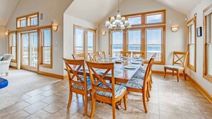 A spacious dining area with a large table set for six, surrounded by wooden chairs with blue cushions. The room has large windows offering a view of the ocean. - Premier Oceanfront Carolina Dreamin’ w/ Pool, Hot Tub, Elevator, Game Room (Avon)