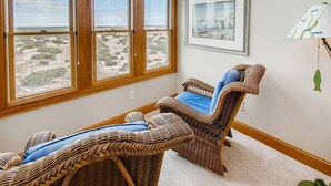 A room with a large window overlooking a desert landscape, two wicker chairs with blue cushions, and a framed picture on the wall. - Premier Oceanfront Carolina Dreamin’ w/ Pool, Hot Tub, Elevator, Game Room (Avon)