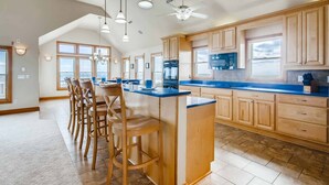 A spacious kitchen with a central island, wooden bar stools, and a large window providing natural light. - Premier Oceanfront Carolina Dreamin’ w/ Pool, Hot Tub, Elevator, Game Room (Avon)
