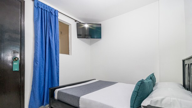 A single bed with a grey and white mattress, a teal pillow, and a grey blanket. A blue curtain is partially drawn, and a TV is mounted on the wall. - Hotel Ayenda Los Tres Pinos (Bogotá)