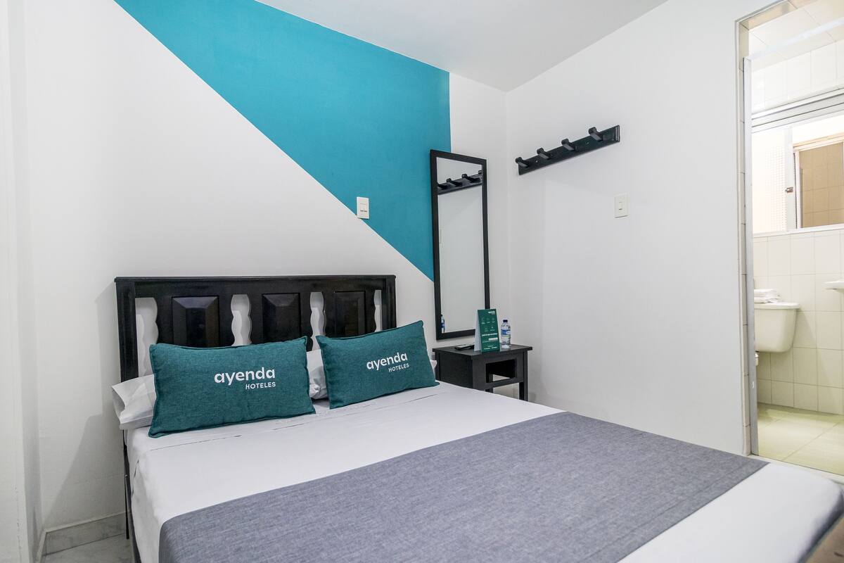 Ein Hotelzimmer mit einem Bett, zwei Kissen mit dem Schriftzug 'ayenda HOTELES', einem Nachttisch mit Spiegel und einem durchgehenden Türspalt sichtbaren Badezimmer.