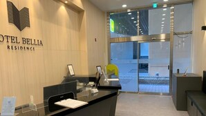 A reception desk at HOTEL BELLIA RESIDENCE with a glass partition, a yellow chair, and a view of the outside through the glass doors. - Hotel BELLIA (Busan)