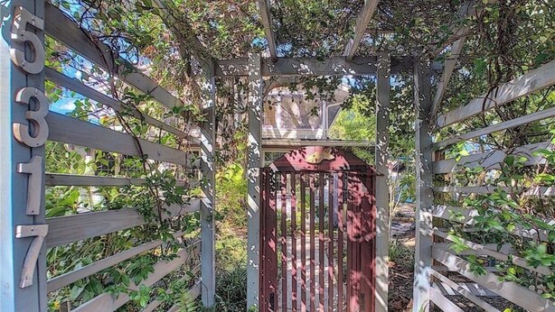 Um portão de madeira com placa de número de metal, cercado por vegetação e um céu aberto.