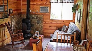 Ein gemütliches Blockhäuschen-Interieur mit Kamin aus Stein, Holzwänden, Fliesenboden und einem Esstisch mit Stühlen.