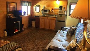 A cozy cabin-style room with a fireplace, a sofa, a dining table, and a kitchen area with a microwave and refrigerator. - One Bedroom Cabin near Mount Rushmore (Rapid City)