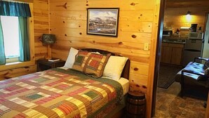 A wooden cabin bedroom with a bed, a nightstand, a picture on the wall, and a view into the kitchen area. - One Bedroom Cabin near Mount Rushmore (Rapid City)