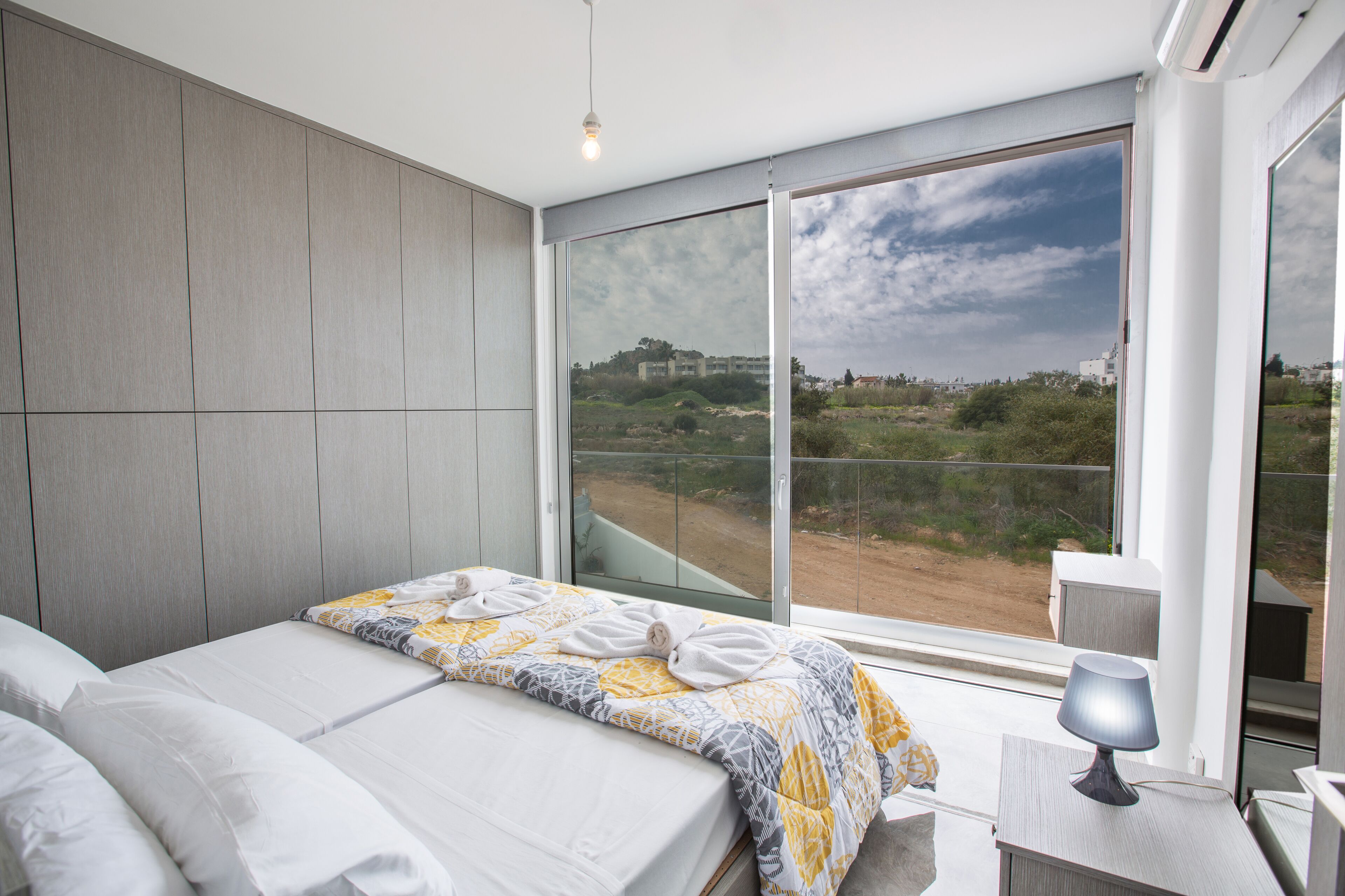 Un dormitorio moderno con una cama grande, una mesita de noche con lámpara y vistas a un paisaje a través de un ventanal corredero.