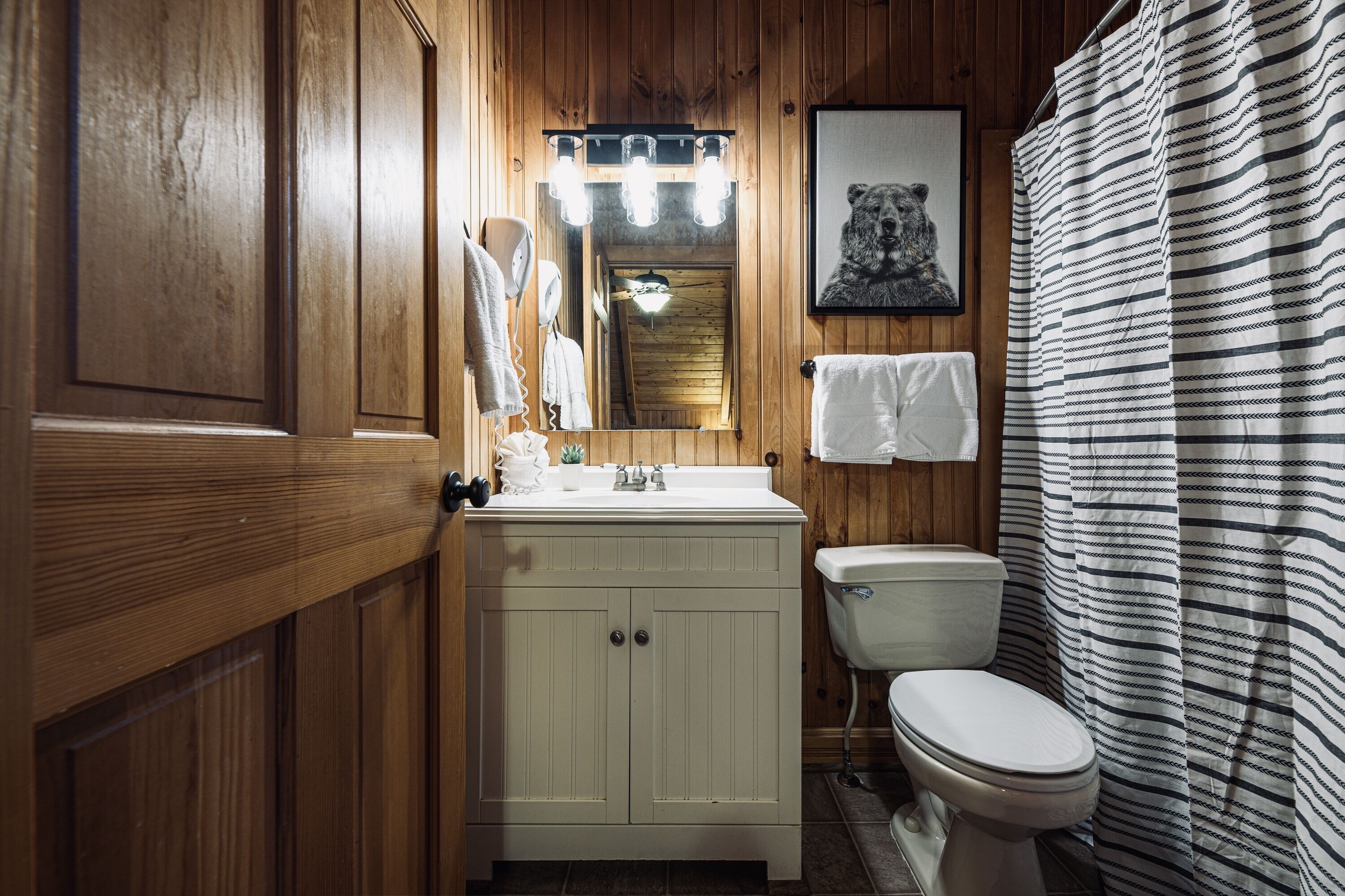 Une salle de bain aux murs lambrissés, avec un lavabo blanc, des toilettes et un rideau de douche à rayures.