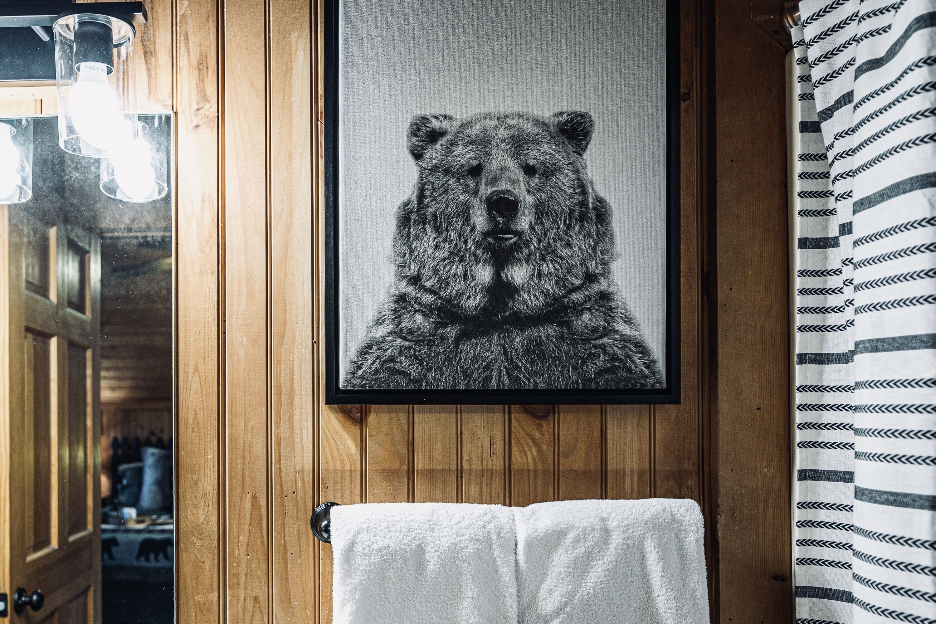Un retrato de un oso en un marco sobre una pared de madera, una toalla blanca colgada de una puerta y una cortina a rayas.