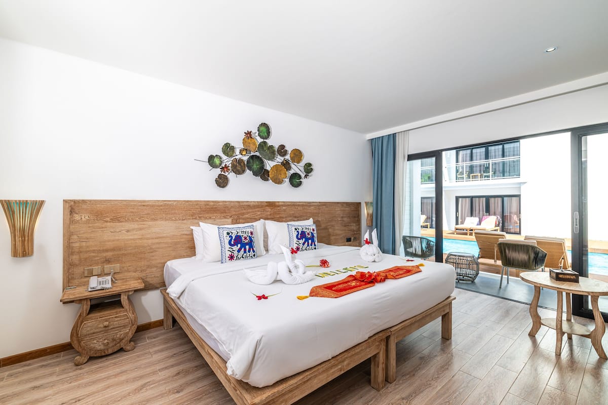 Ein Hotelzimmer mit Holz-Kopfteil, einem Bett mit weißen Laken, Dekokissen, einem Wandbild, einem Nachttisch mit Telefon und Blick auf den Pool durch große Fenster.