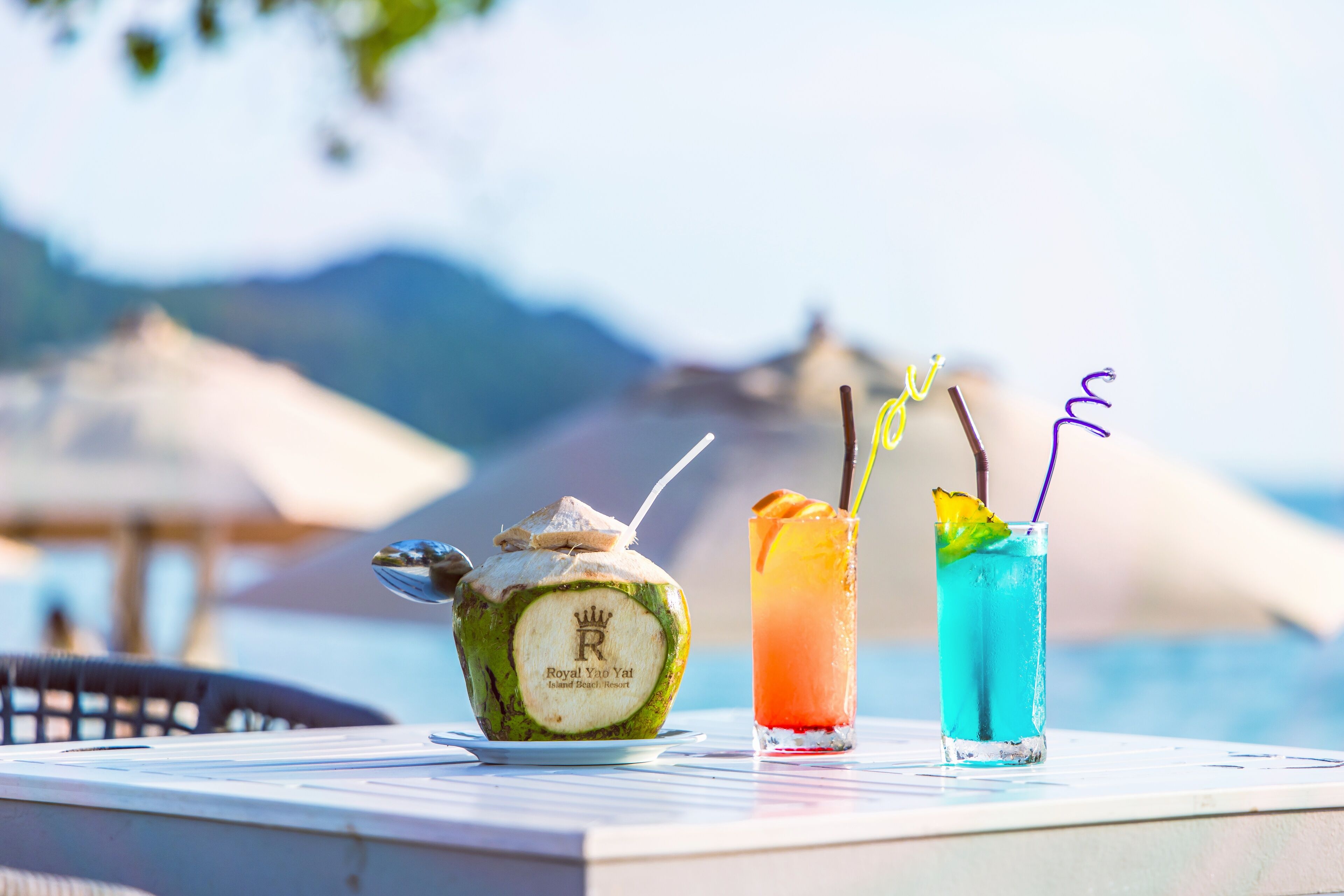 Una mesa con un refresco de coco y dos cócteles, con una vista panorámica del mar y las montañas al fondo.