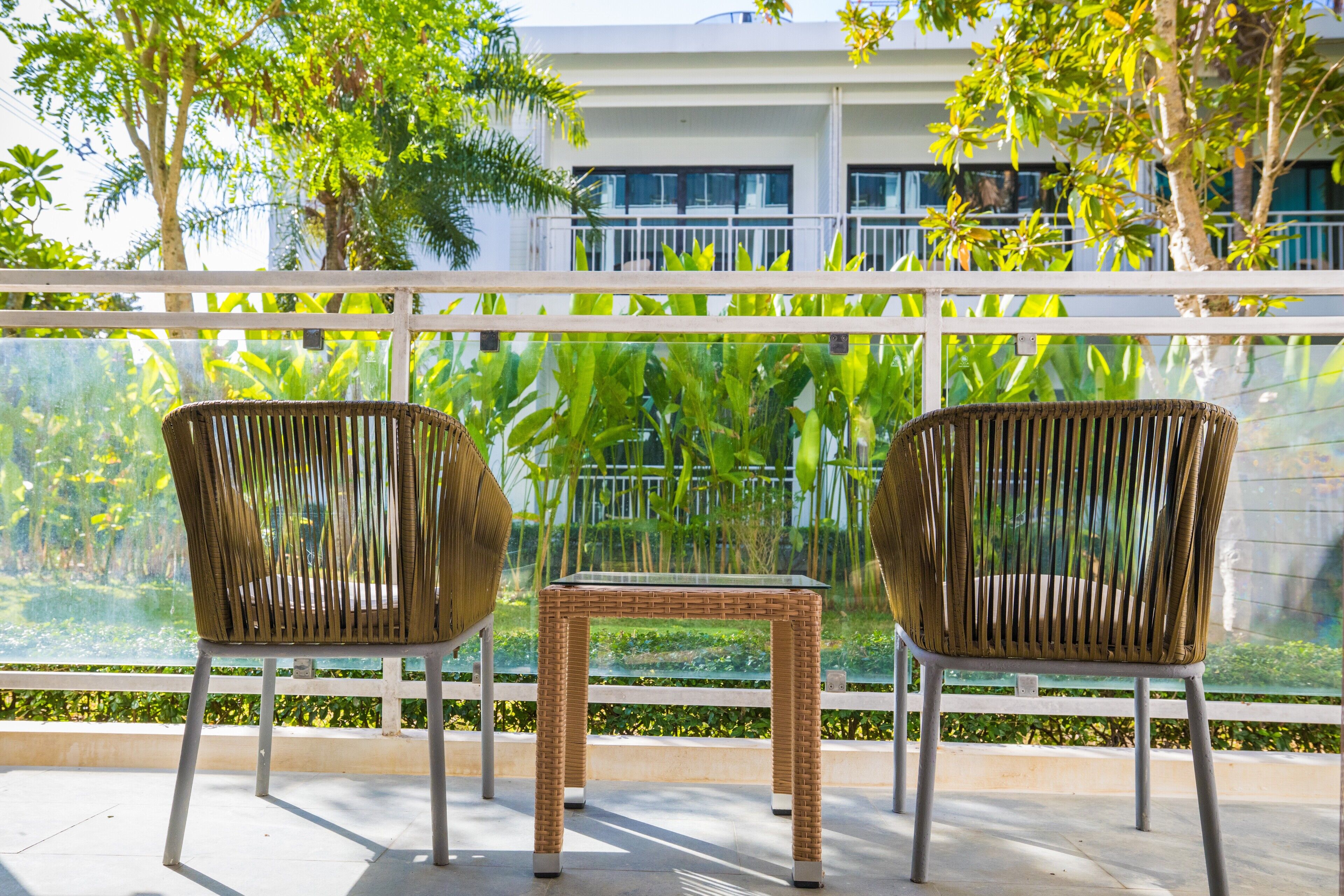 Deux chaises en osier et une petite table sur un balcon, avec vue sur la verdure et un bâtiment en arrière-plan.
