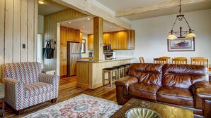 A spacious living room with a brown leather sofa, patterned armchair, and a coffee table. A kitchen with wooden cabinets and a dining area with wooden chairs are visible in the background. - Best Location - True Hike In/Out TH w/Private HotTub and Slope Views-Gas Fireplace-W/D in Unit (Steamboat Springs)