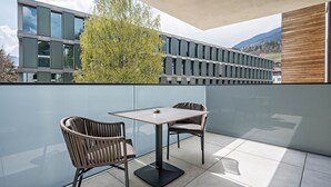 Un balcón con mesa y dos sillas, una pared de cristal y vistas a un edificio y un árbol.