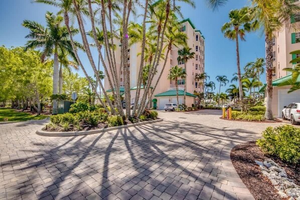 A paved courtyard with palm trees, a building with a green roof, and parked cars.