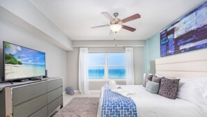 Ein Schlafzimmer mit Bett, Deckenventilator, Fernseher auf einer Kommode, Fenster mit Vorhängen und einem Bild an der Wand.